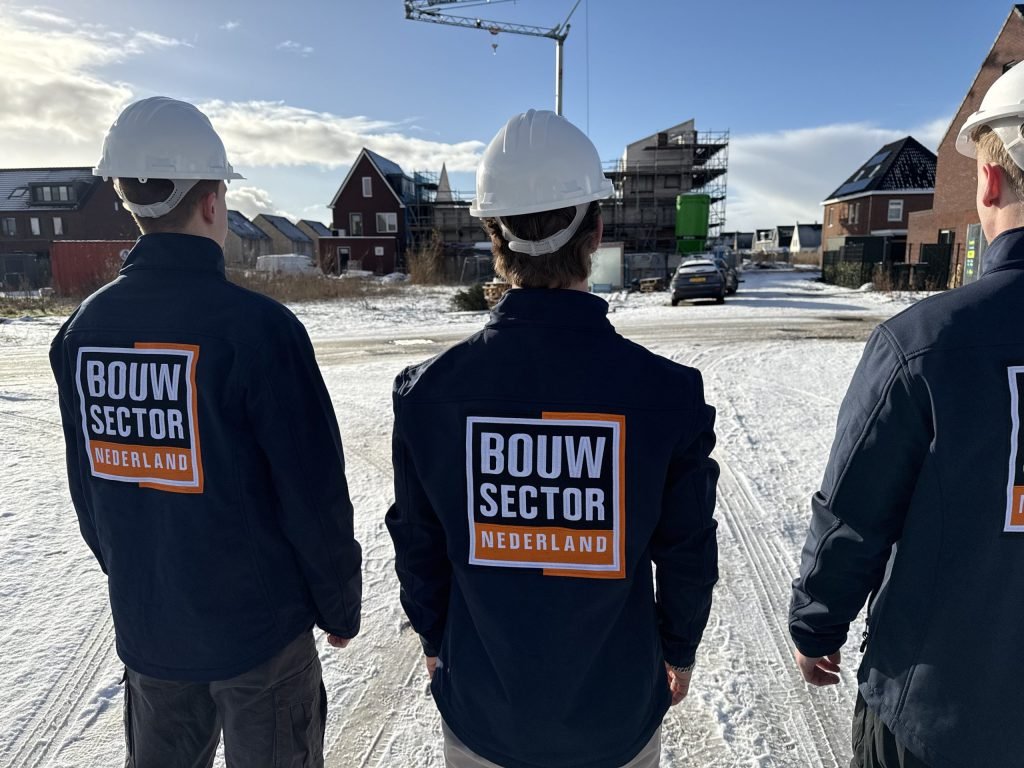 Drie vakmensen van Bouwsector Nederland met witte helm en logo op de rug bij huizen in aanbouw